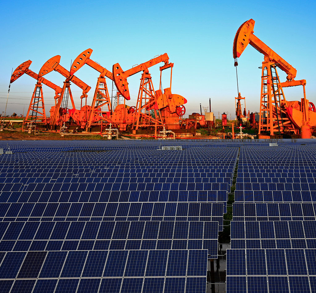 A field of solar panels with oil pumps in the background.