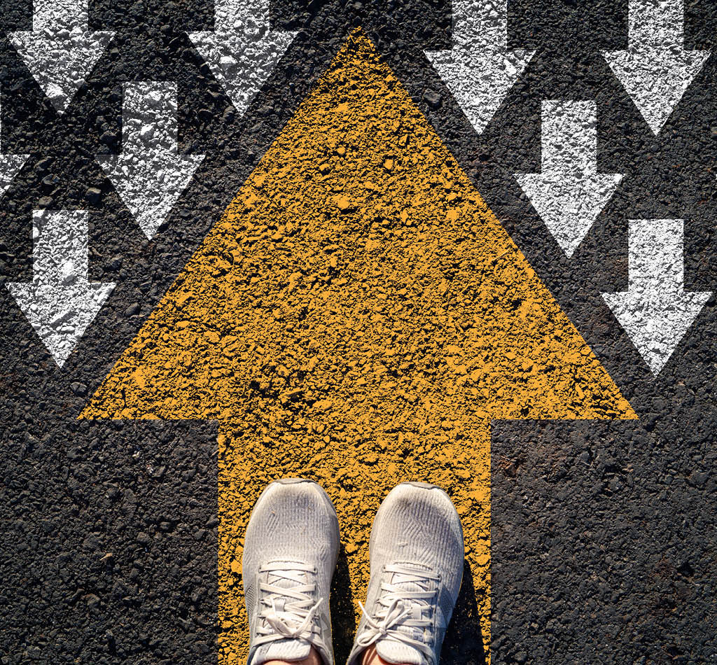 Top view of shoes standing on a bold yellow arrow pointing up against a field of white arrows pointing down.