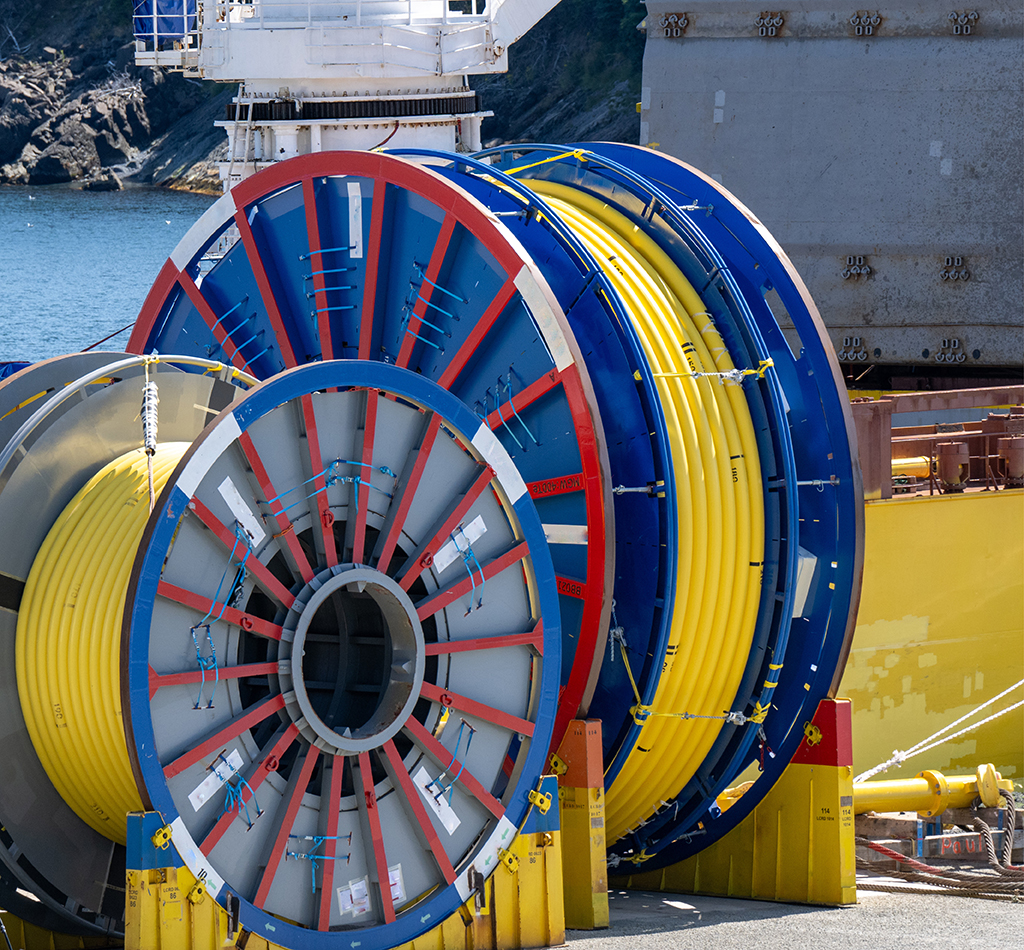 Bulk sub-sea industrial glass fiber optic cable on a metal spool on a ship's stand. The yellow data line is coiled around a black reel in a storage yard.